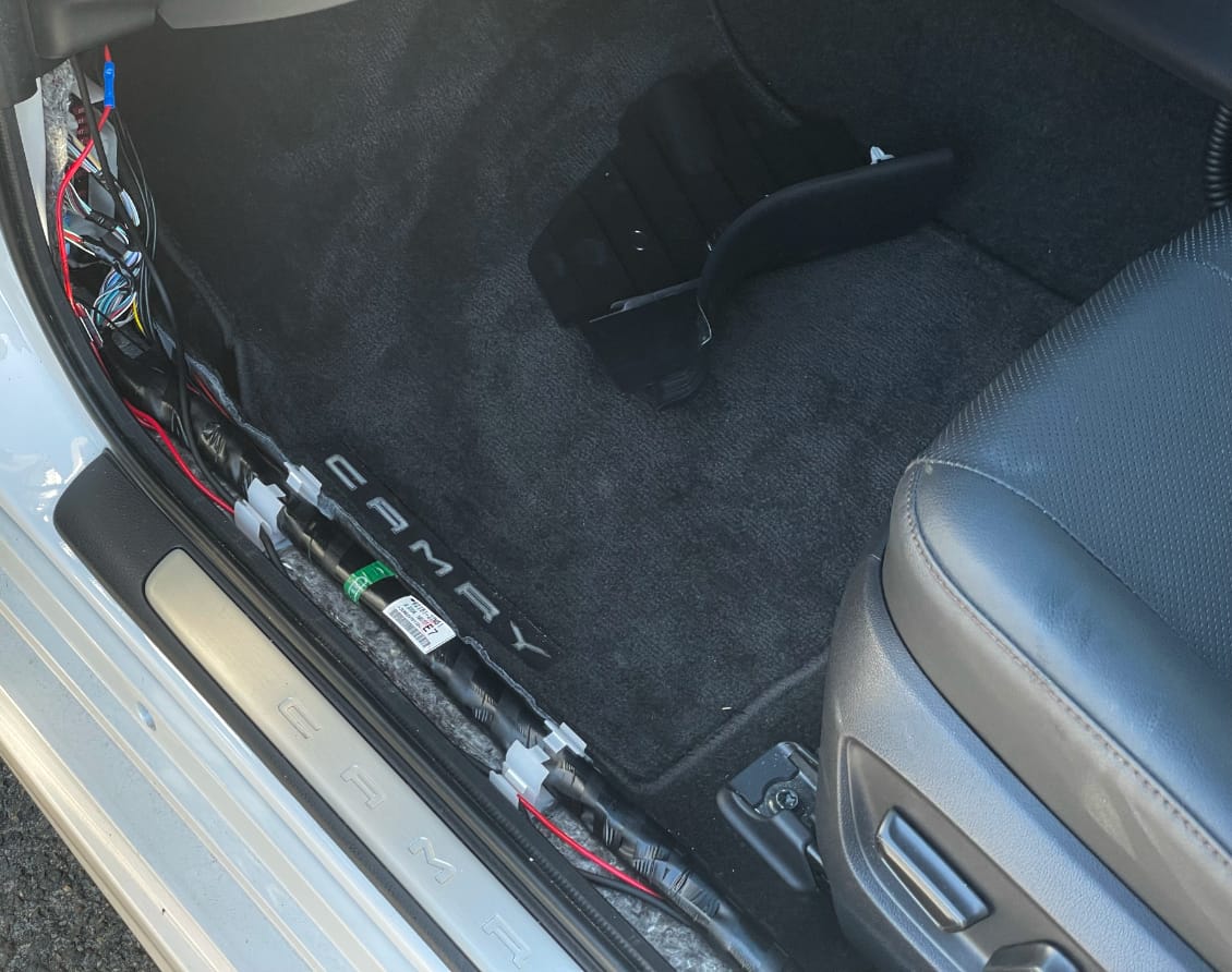 The bottom of the front passenger's door frame. Two pieces of trim have been removed, exposing a number of cables. One of those pieces of trim is seen sitting in the passenger's footwell.